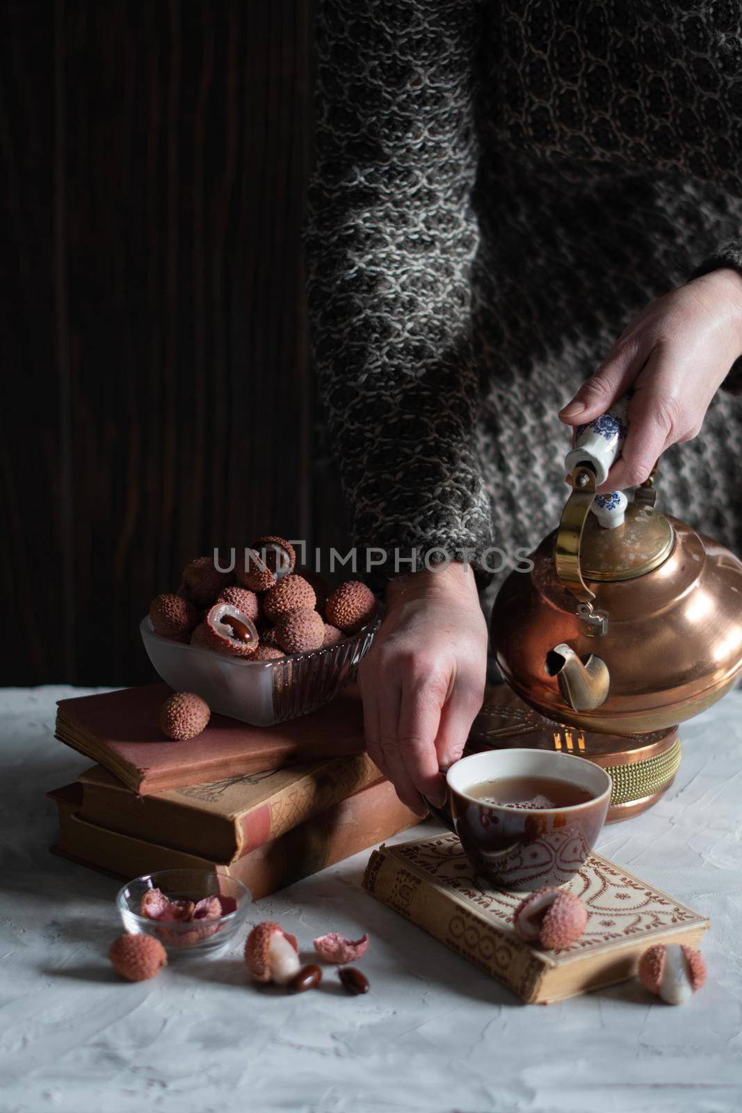 A woman pours tea from a copper teapot into a cup against the background of a stack of books and a cup with lychee, homely cozy tea drinking and reading,. High quality photo