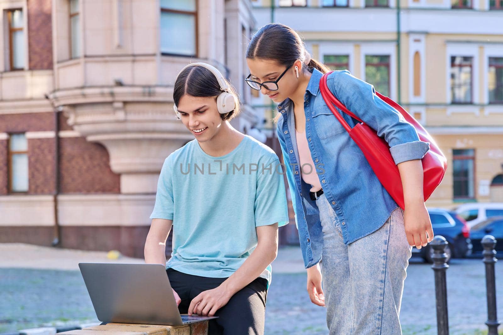 Teenage male and female using laptop for study, leisure, outdoor on city street. Guy and girl teenagers looking together at aptop screen. Lifestyle, technology, youth, education, city life concept