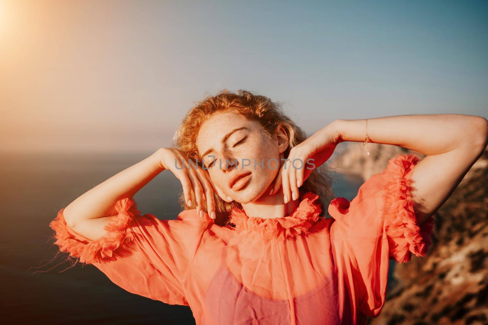 Side view a Young beautiful sensual woman in a red long dress posing on a volcanic rock high above the sea during sunset. Girl on the nature on overcast sky background. Fashion photo