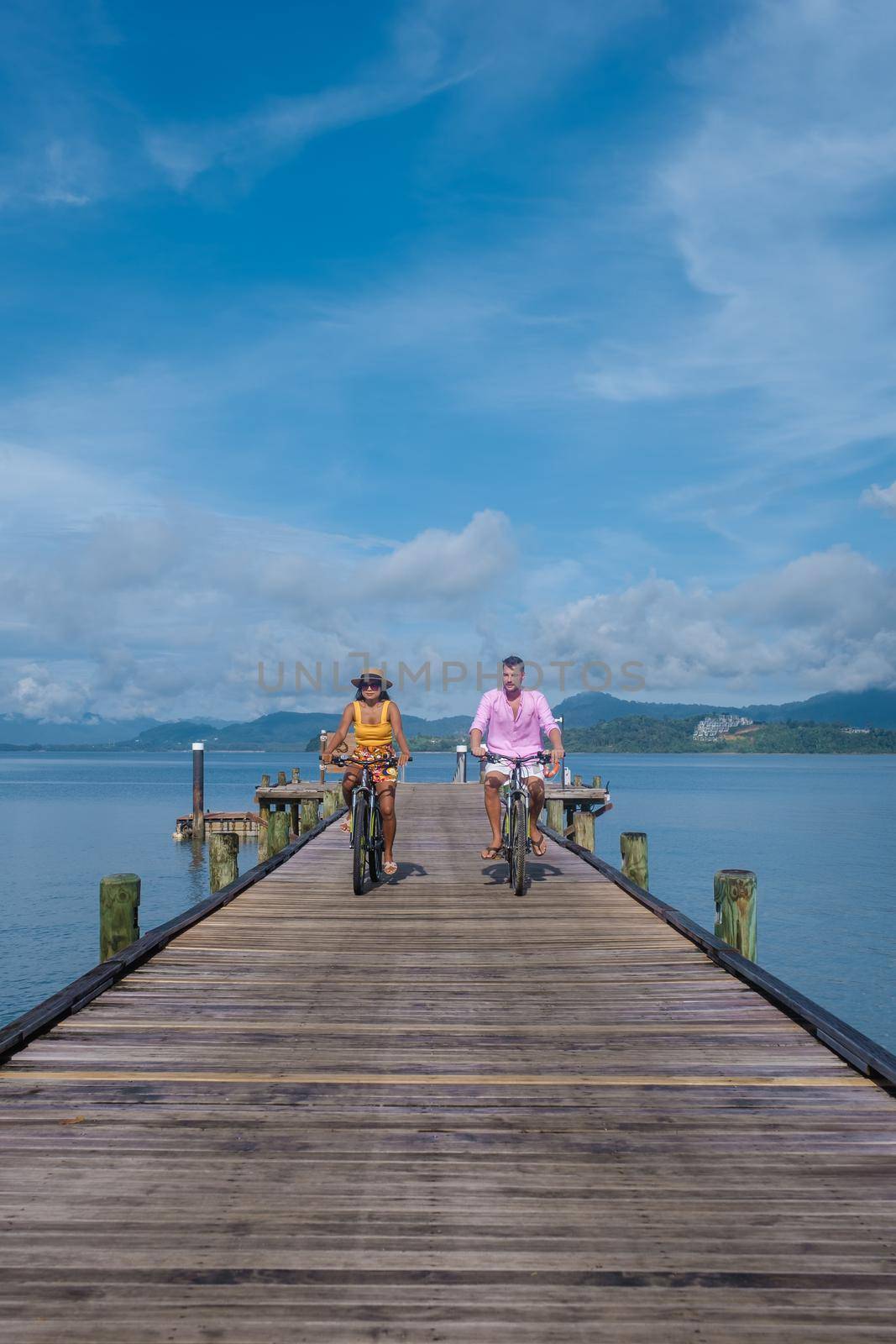 men and woman on a bicycle, couple men and woman on a tropical island with wooden pier jetty in Thailand Phuket Naka Island by fokkebok