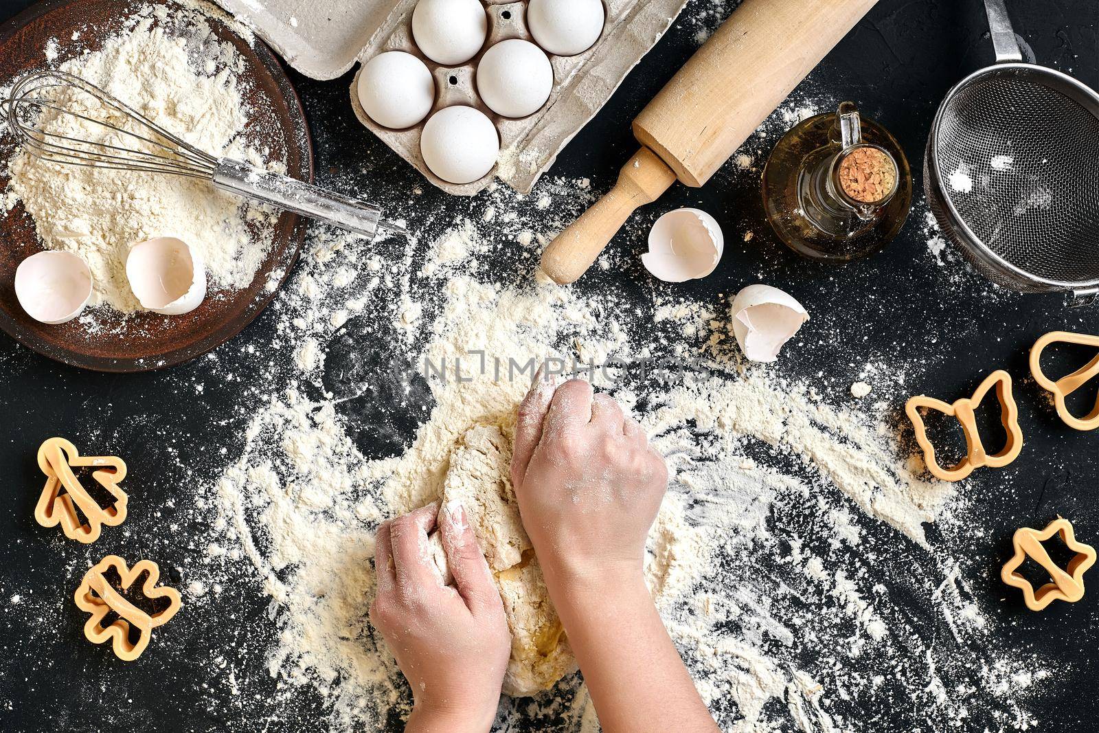 Woman's hands knead dough on table with flour, eggs and ingredients on black table. Top view. Still life. Flat lay