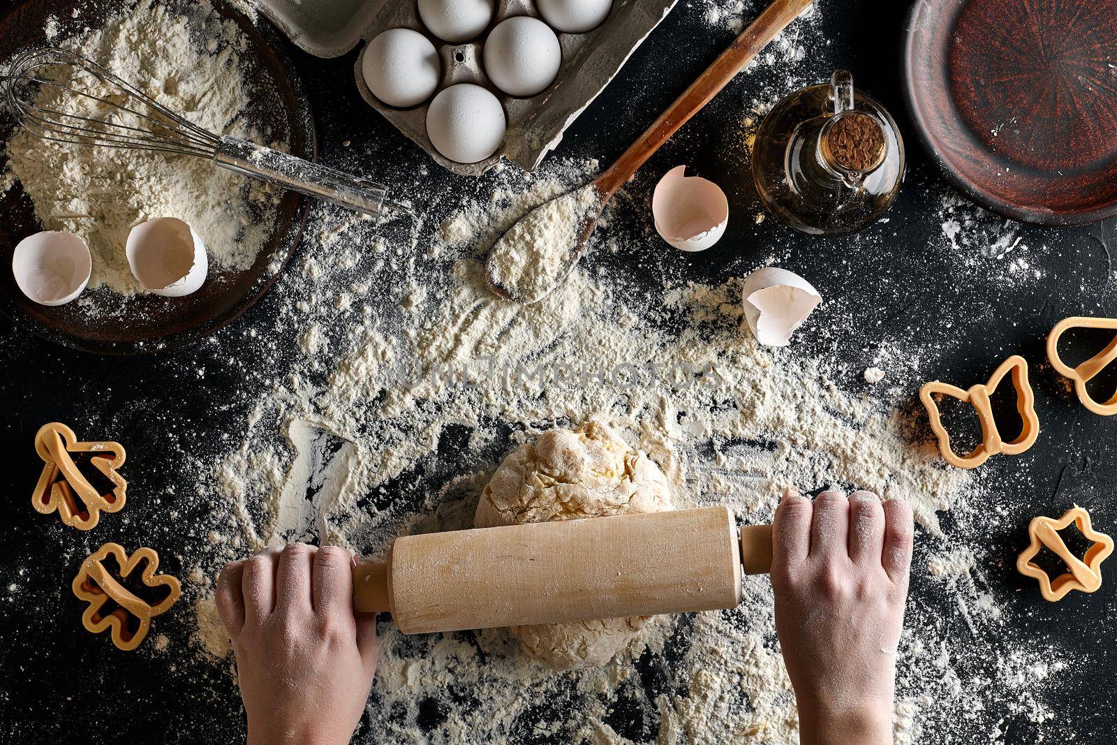 Female hands rolling dough with a rolling pin on a black table. Ingredients for the dough: flour, oil and eggs. Top view. Still life. Flat lay