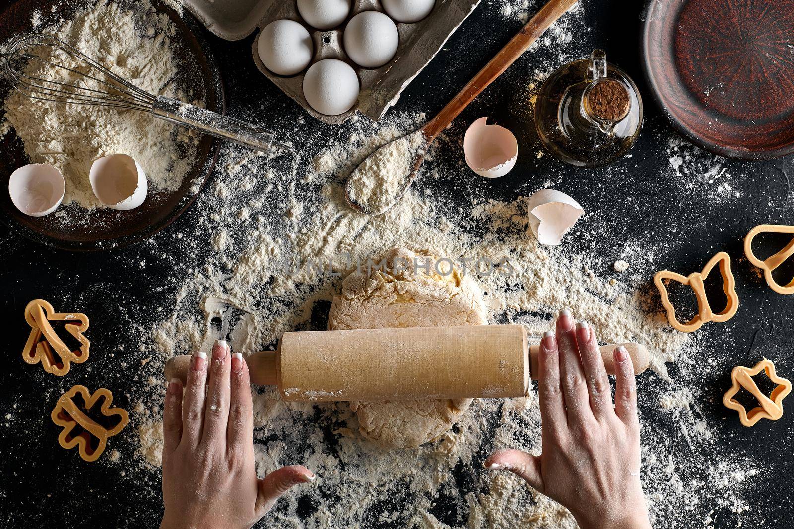 Female hands rolling dough with a rolling pin on a black table. Ingredients for the dough: flour, oil and eggs. Top view. Still life. Flat lay