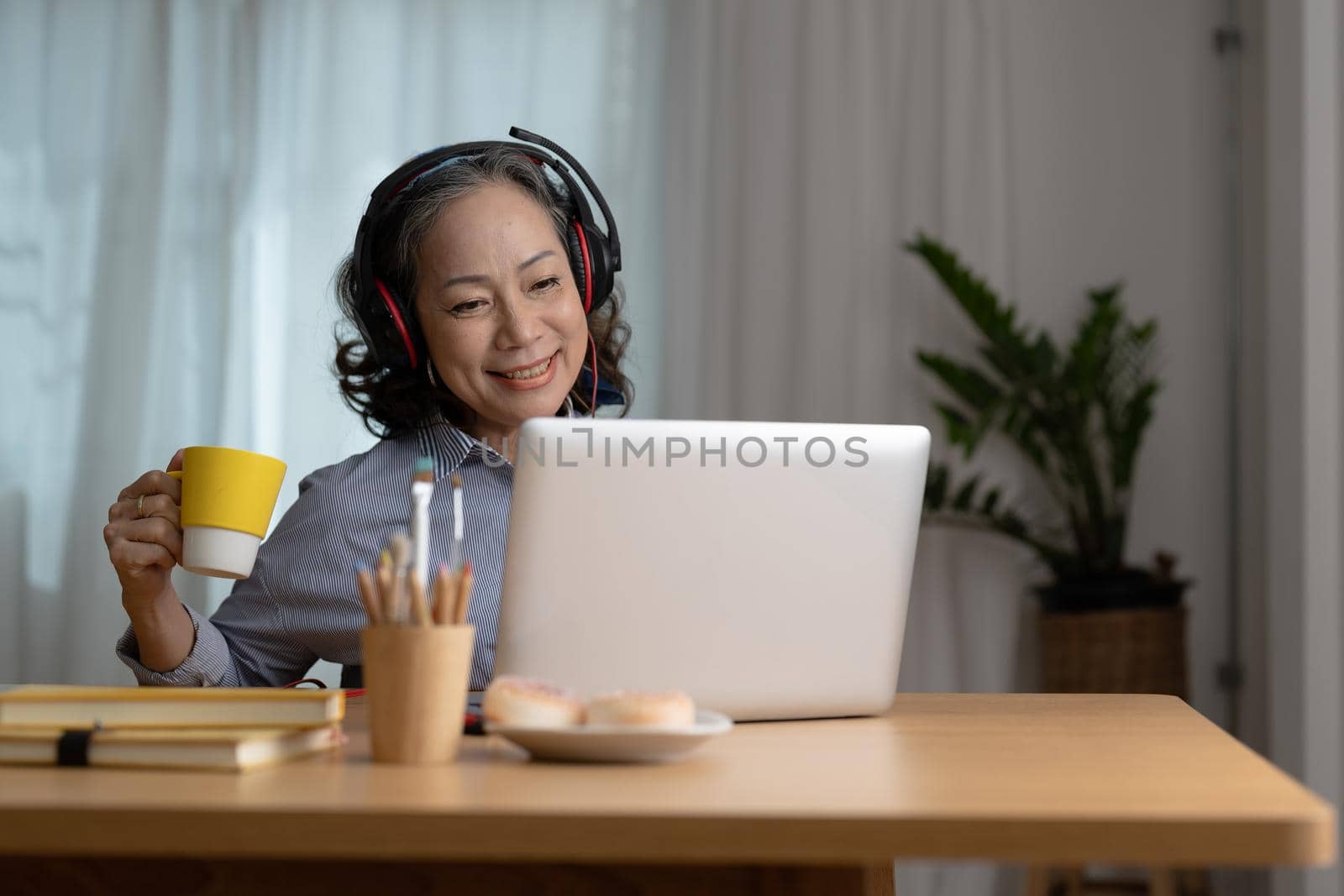 Happy senior asian woman wearing headphones listens songs on laptop in living room at cozy home.