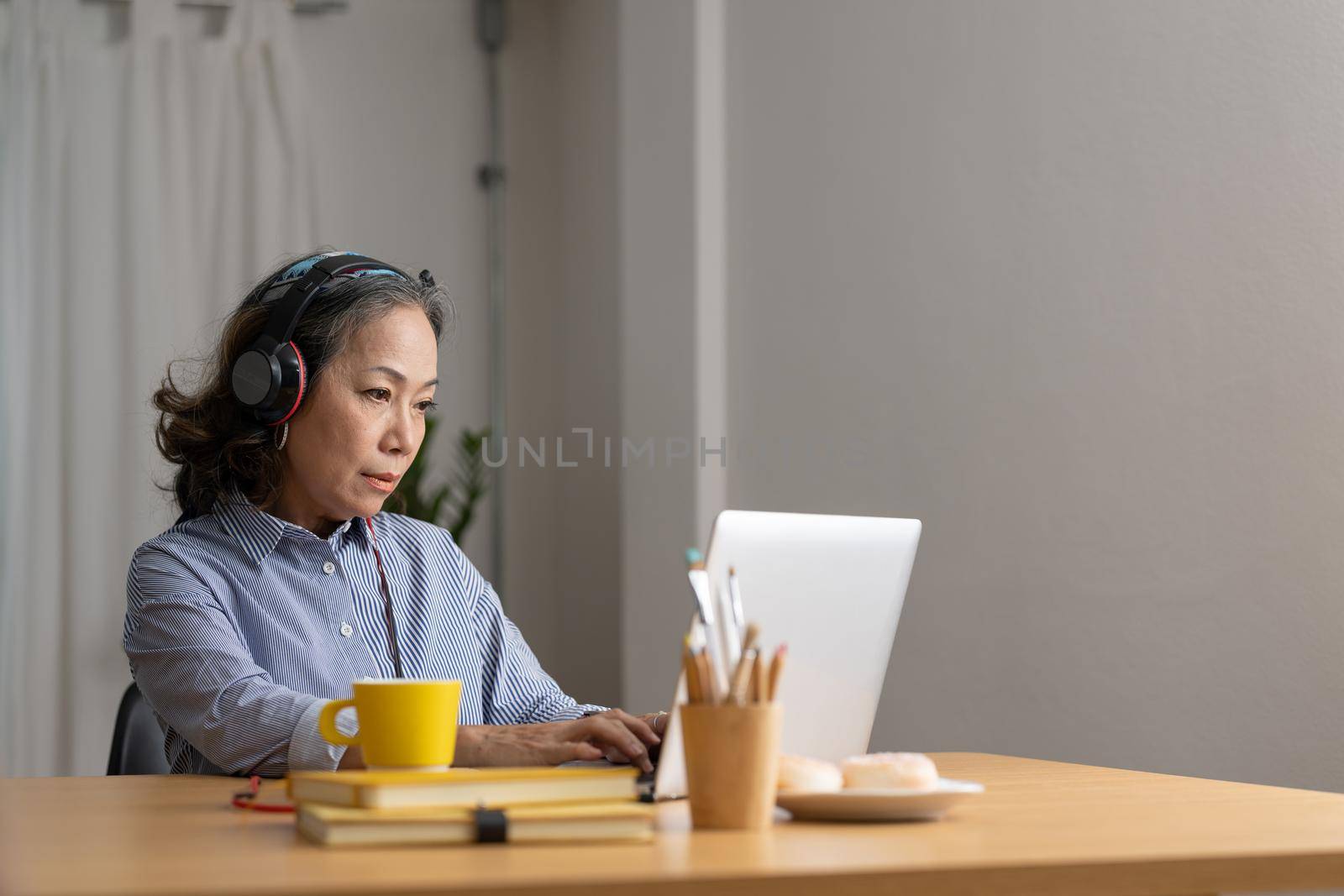 Mature asian woman wearing headphones using laptop, making video call, sitting at table in kitchen, senior teacher mentor wearing glasses engaged online conference, recording webinar, teaching.