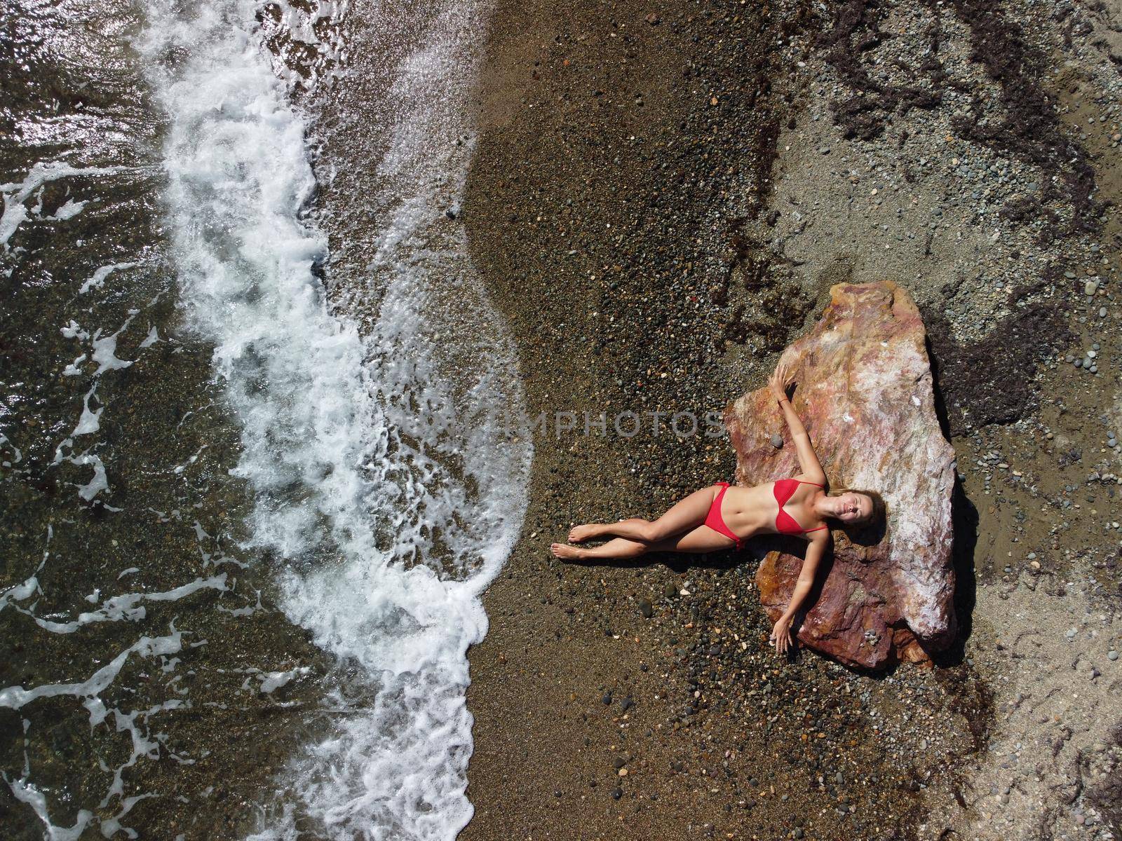 Young sensual blonde woman sitting on the rock near water at sea and enjoys the sea waves on background of two volcanic rocks, like in Iceland. Dreams holidays and weekend vacation in summer time