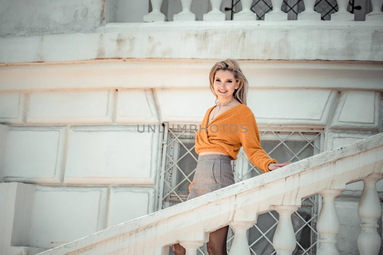 A middle-aged woman looks like a good blonde with curly beautiful hair and makeup on the background of the building. She is wearing a yellow sweater