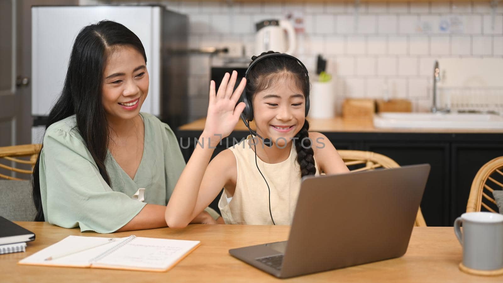 Cheerful asian girl in headphones talking to teacher while studying online via laptop at home with happy mom sitting nearby and giving support by prathanchorruangsak