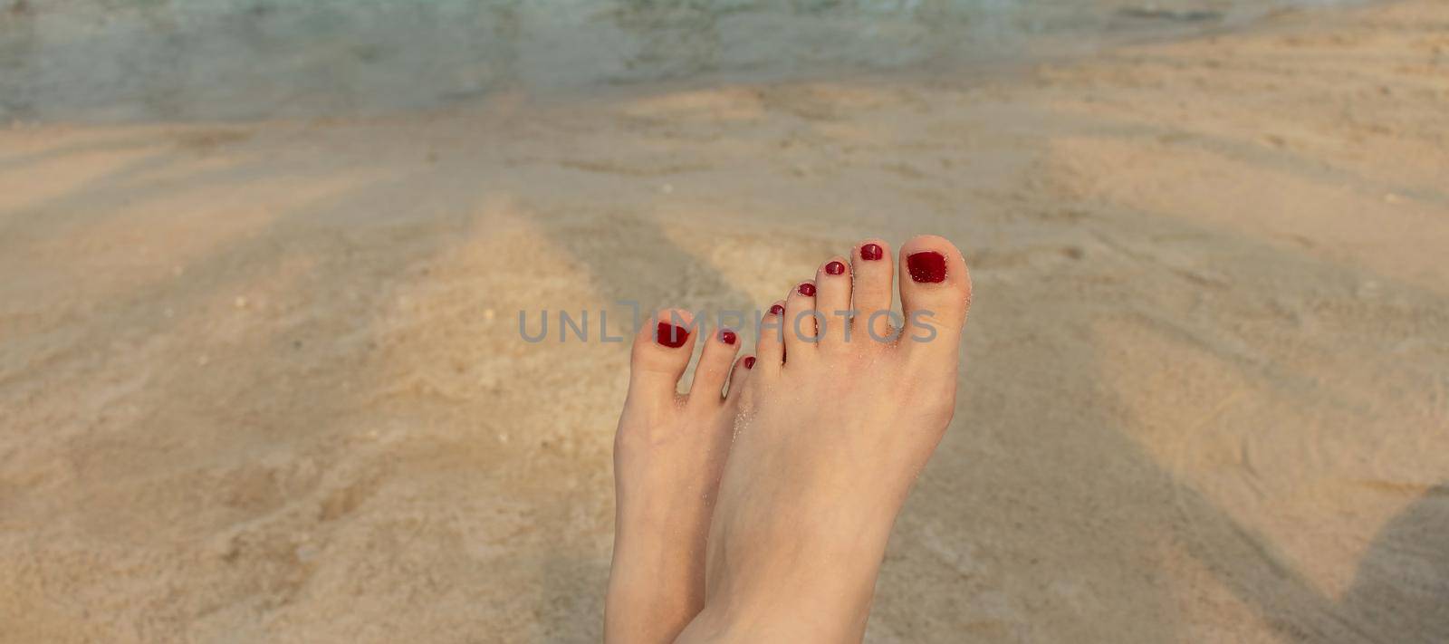 banner with beautiful female feet with red pedicure on the background of blue water and sand on the beach. the concept of relaxation by the sea on a bright, hot and sunny day by Leoschka