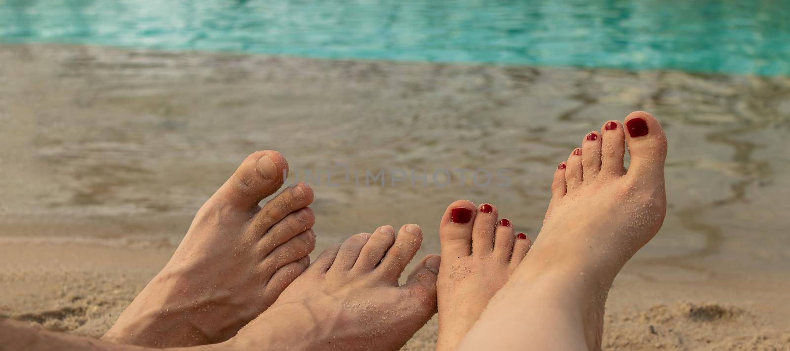 banner with male and female feet lie on the sand against the background of blue water. two pairs of legs. concept of relaxation by the sea on a bright, hot and sunny day on the beach by Leoschka