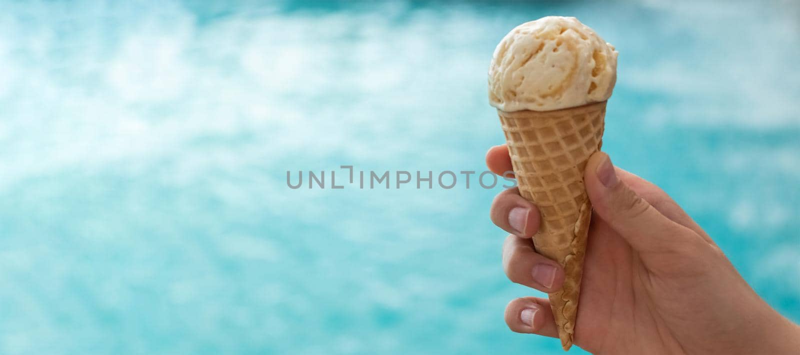 banner with young hand holds an ice cream cone on the background of the blue water of the sea. the concept of relaxation by the sea on a bright, hot and sunny day. soft focus