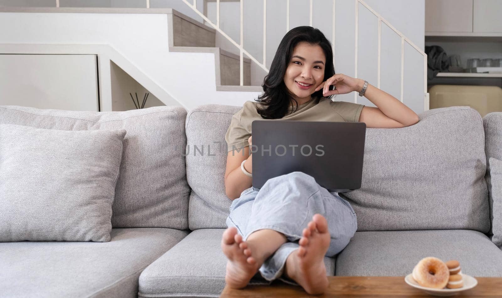 Beautiful smiling Asian woman looking at camera relaxing on leather sofa at home. work on a laptop computer.