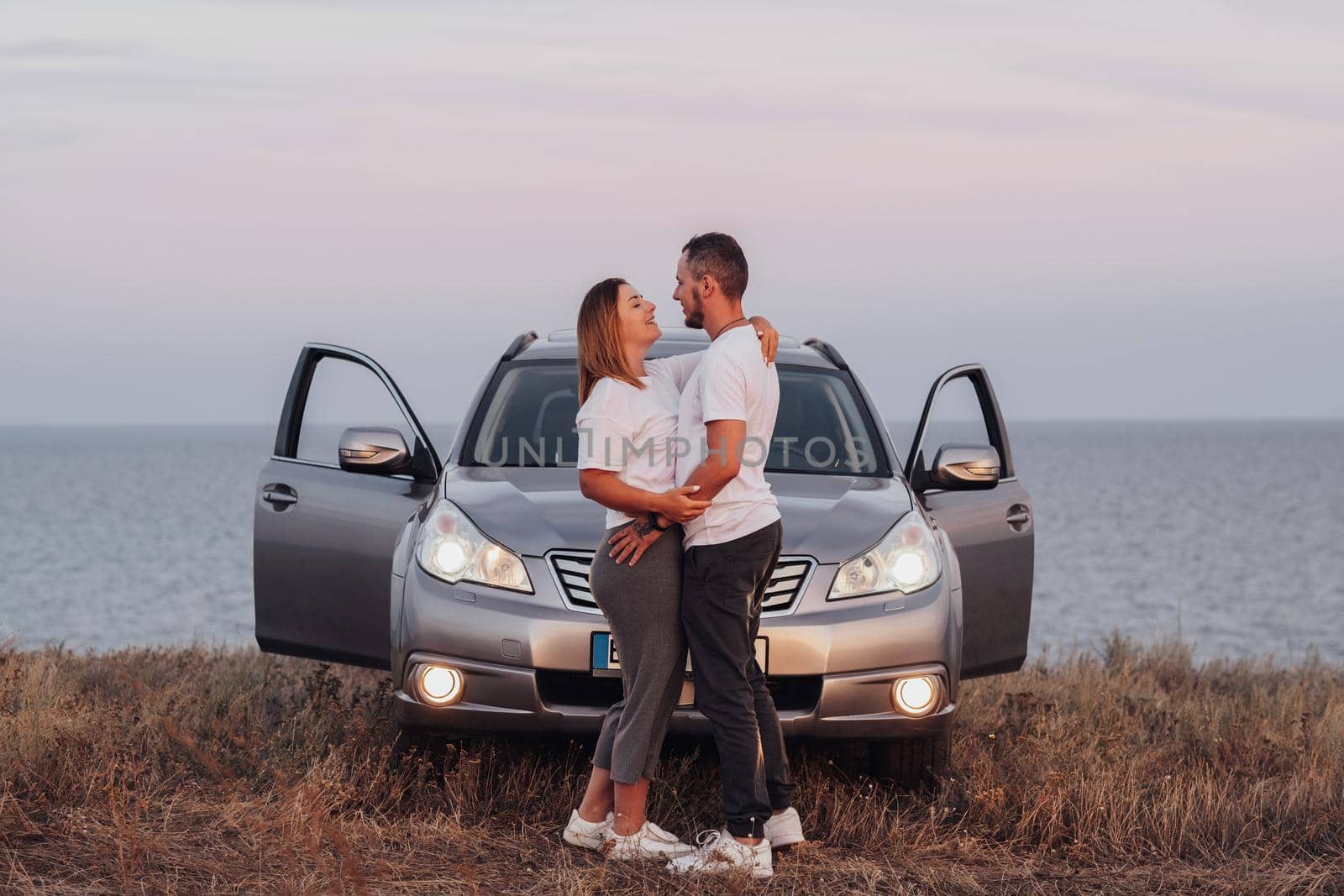 Man and Woman Hugging in Front of Their SUV Car, Young Couple Standing Together on Background of Sea, Road Trip Concept