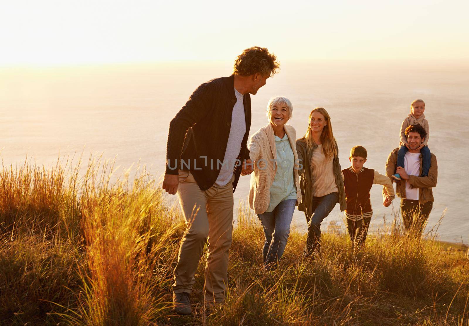 A multi-generational family walking up a grassy hill together at sunset with the ocean in the background.