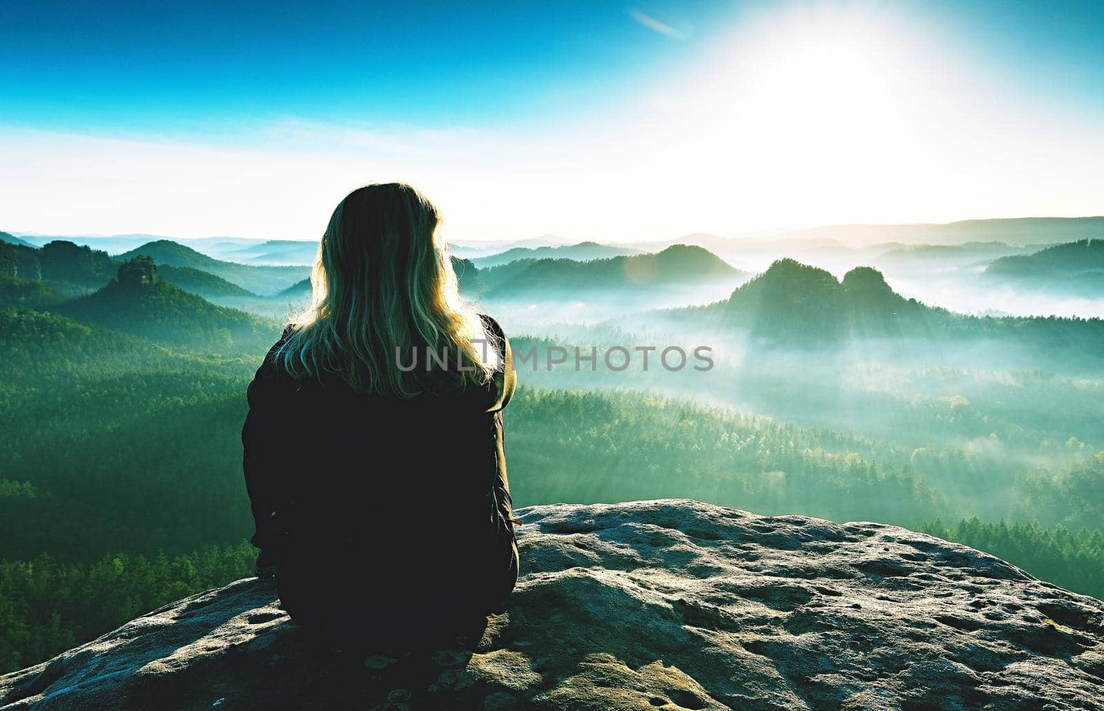 Slim fair hair girl with warm jacket sit on rocky mountain top against bright blue morning sky enjoying foggy mountain range panorama. Tourism, traveling and healthy lifestyle concept.