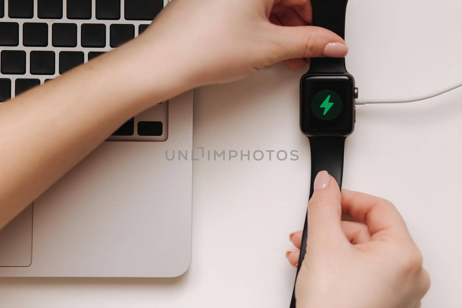 Female hand put smart watch on wireless charging with on-screen charging indicator. Work place near at the laptop. Top view.