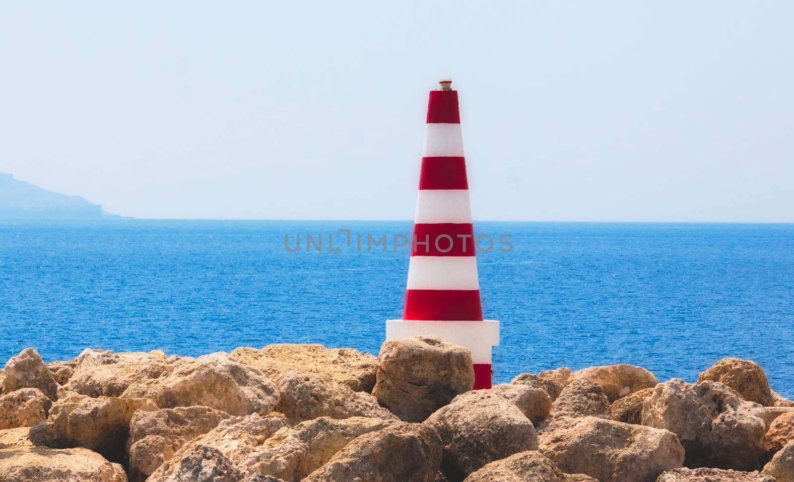 Red and white striped sea safety beacon fixed on the rocks in port with the water horizon in the background on a clear sunny day with a blue sky