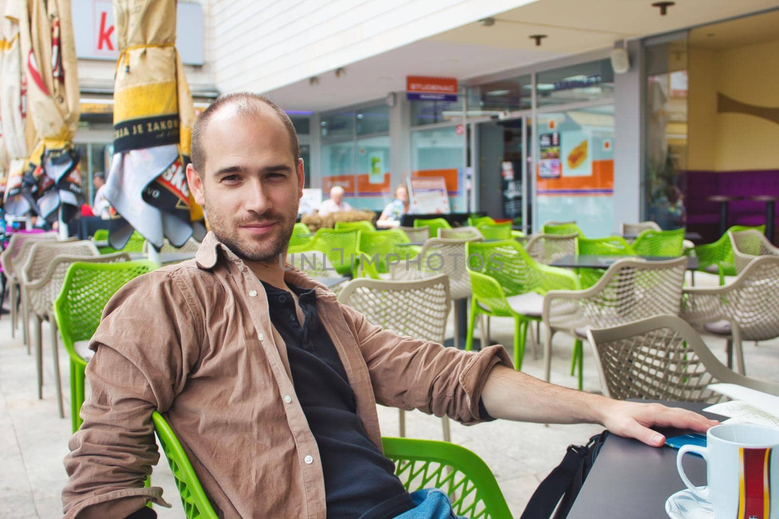 Adult man with casual clothing sitting at a table outside a coffee shop smiling and looking at the camera