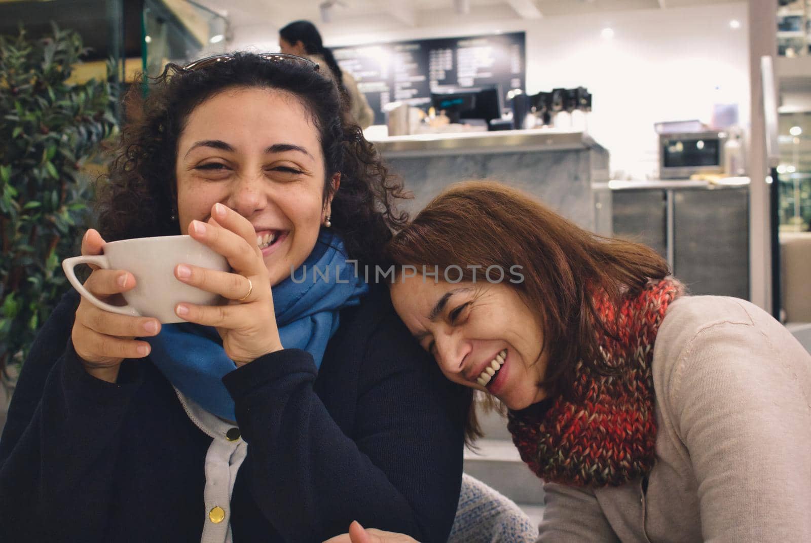 Two women friends (mother and daughter) enjoying a joke and a chat and a cup of coffee or tea, laughing and smiling in a cafe