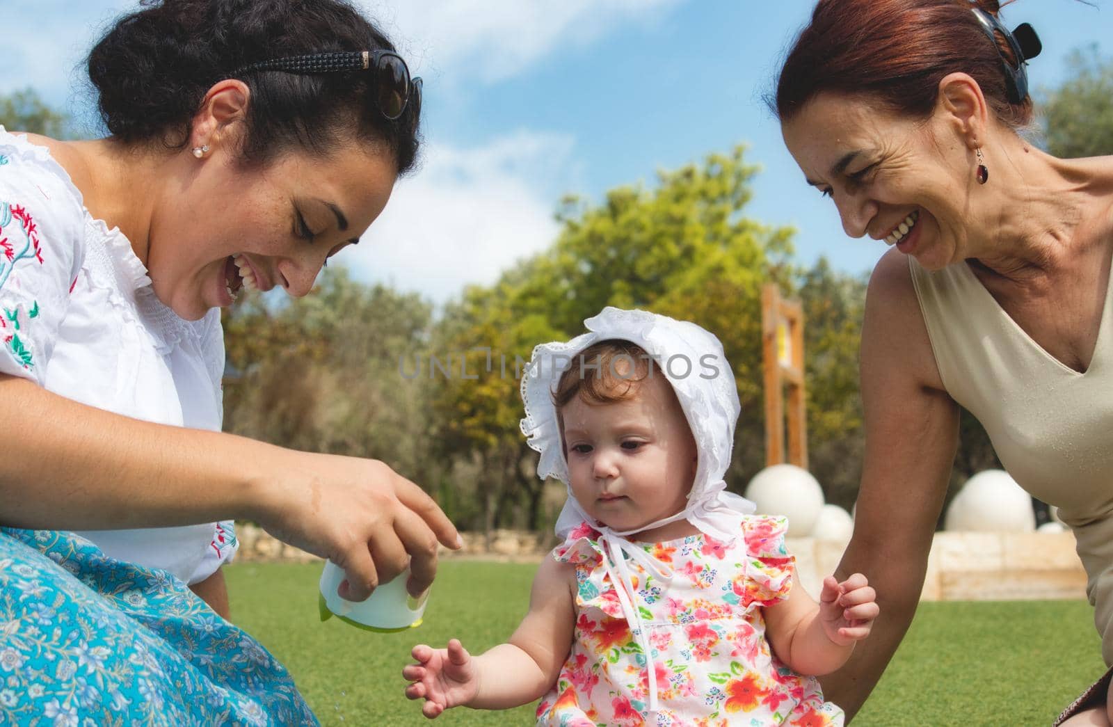 A happy family of 3 generations, baby, mother and grandmother sitting on the grass and playing outdoors in a public park