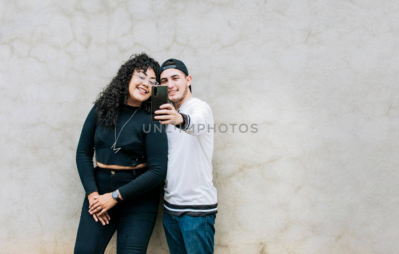 Young couple taking a selfie near a wall with copy space, Young friends taking a selfie leaning against a wall, Young happy couple taking a selfie leaning against a wall