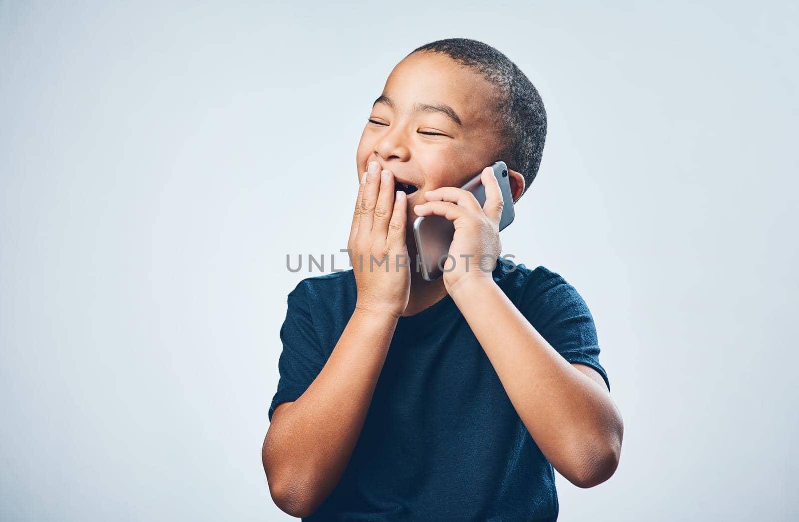 A call from grandma always gives him the giggles. Studio shot of a cute little boy looking amazed while using a smartphone against a grey background. by YuriArcurs