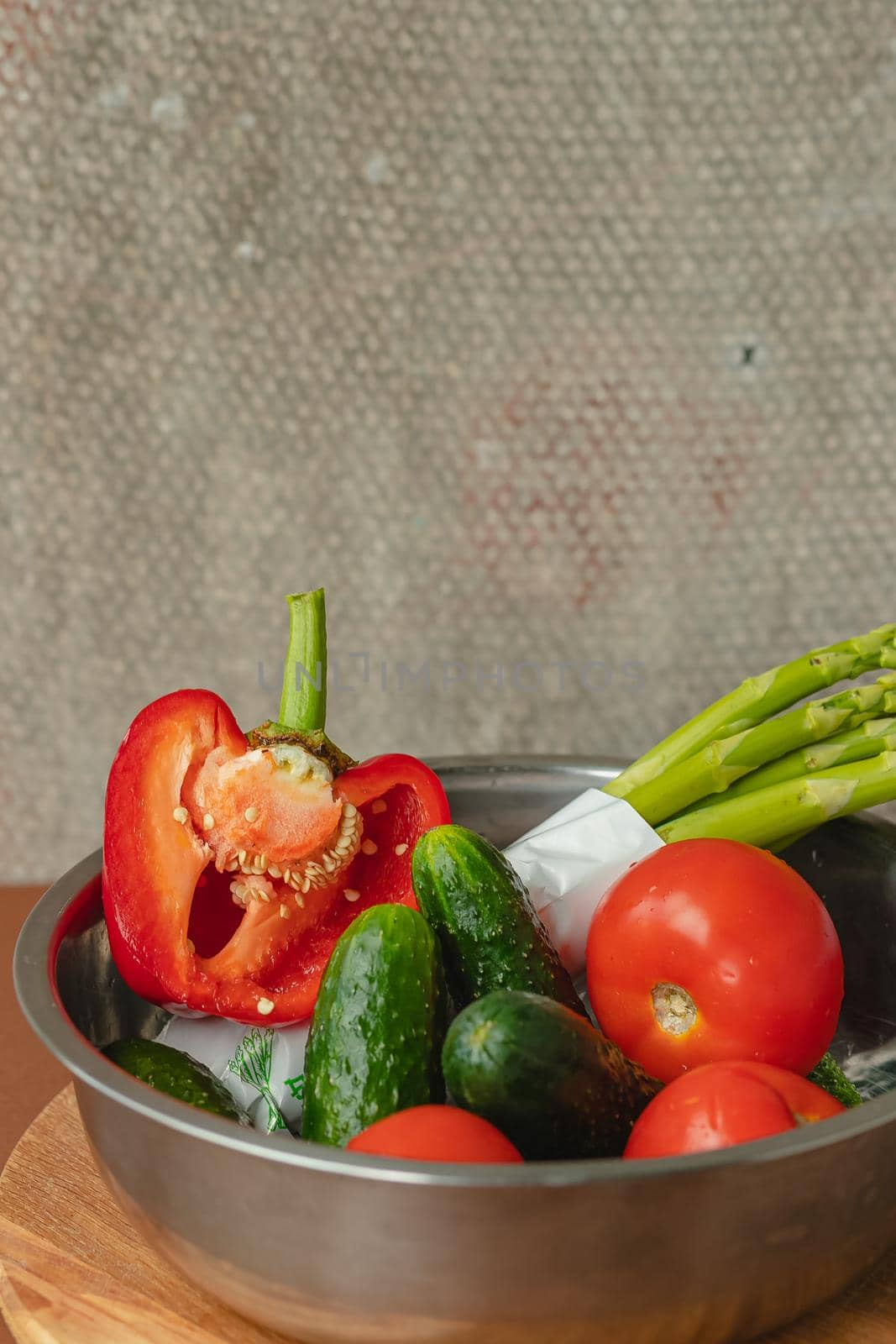 Vegetables lie in a metal bowl tomatoes, asparagus, cucumbers, red bell peppers . on a wooden board and brown background. back gray background. place for text.