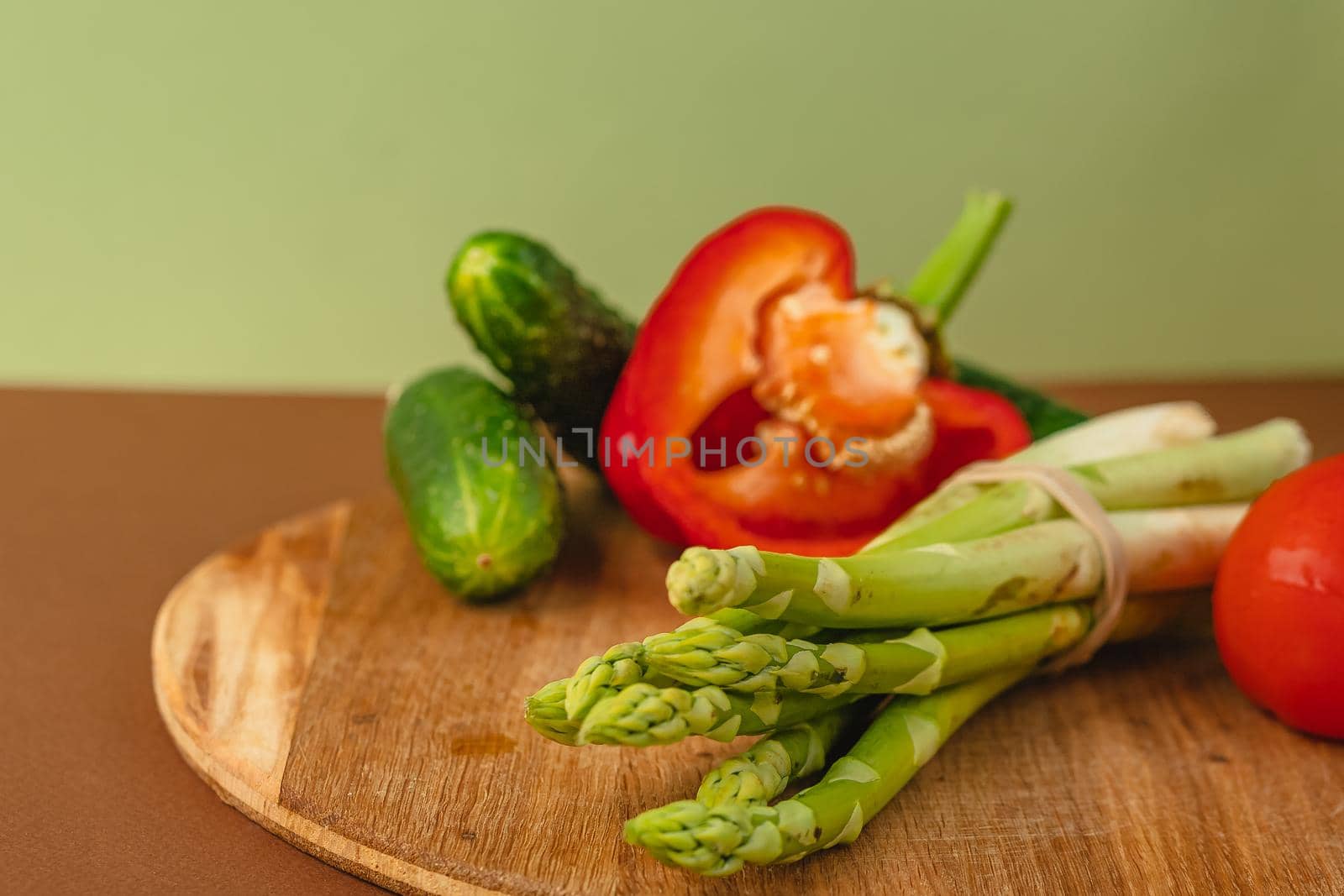 Vegetables lie on a wooden board: tomatoes, asparagus, cucumbers, red bell peppers. brown, light green background. place for text