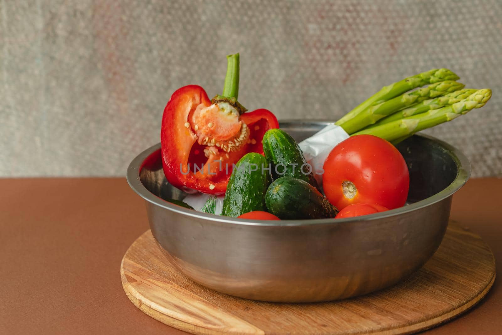 Vegetables lie in a metal bowl tomatoes, asparagus, cucumbers, red bell peppers . on a wooden board and brown background. back gray background. place for text.