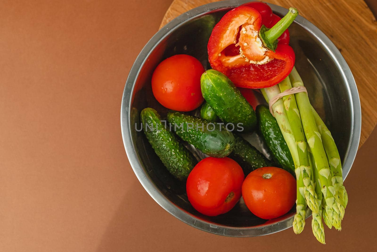 Vegetables lie in a metal bowl: tomatoes, asparagus, cucumbers, red bell peppers. on a wooden board and brown background. place for text. view from above.