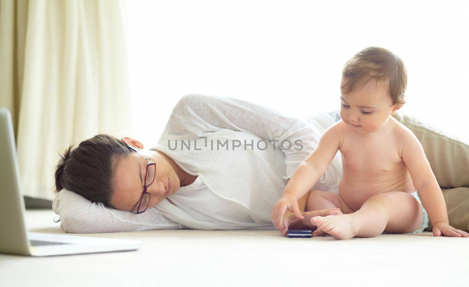 An exhausted young mother lying asleep on the floor with her baby sitting next to her.