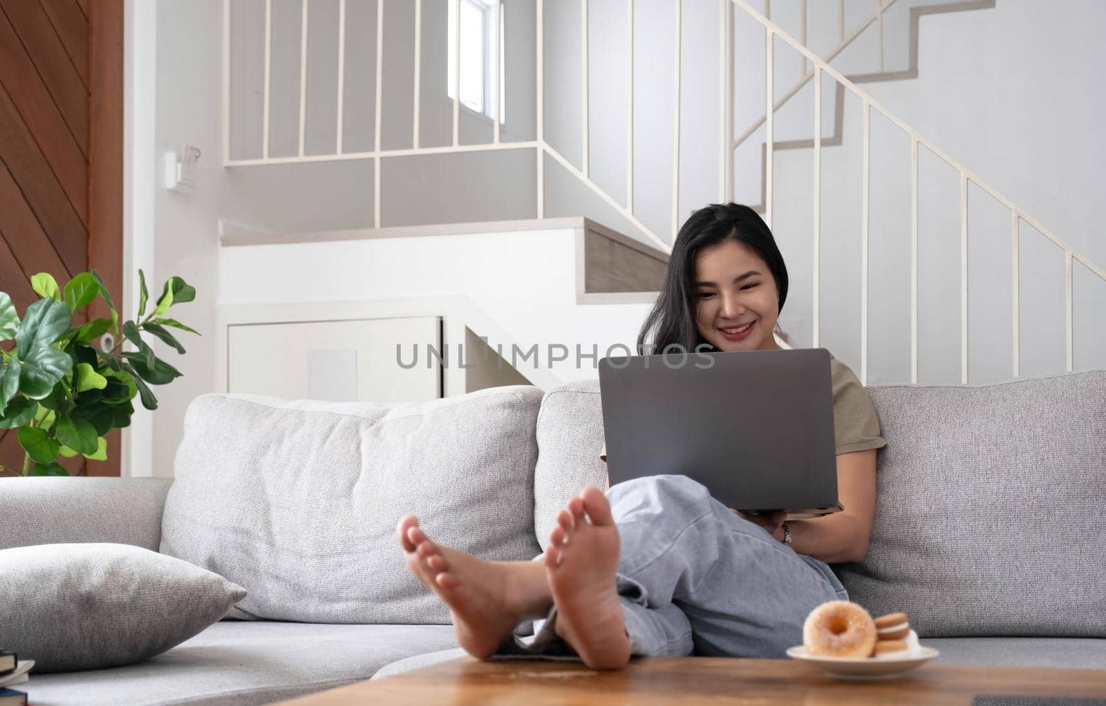 Young business freelance Asian woman working on laptop checking social media while lying on the sofa when relax in living room at home..