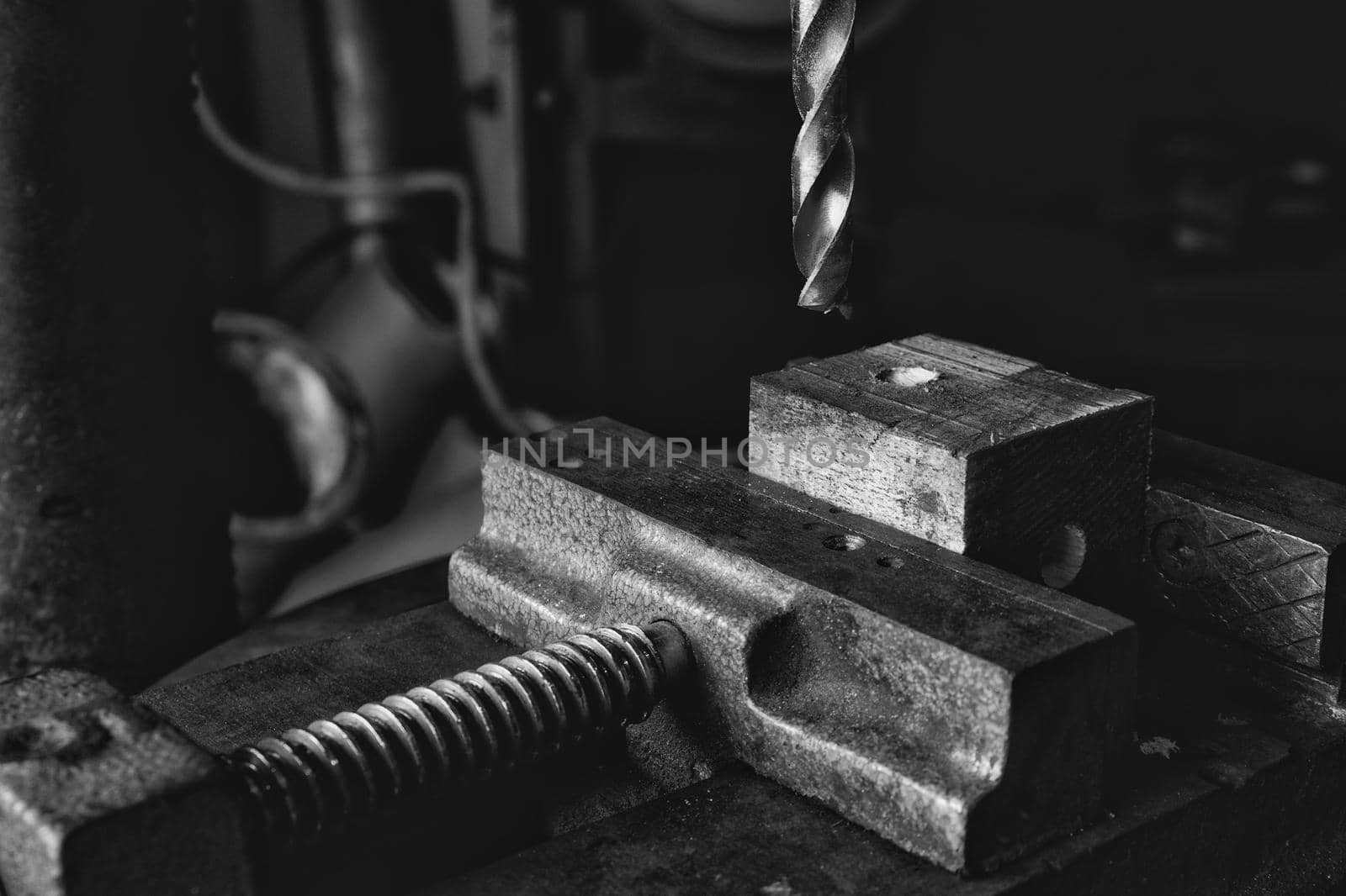 Black and white photo of drill bit coming down onto a piece of wood held in a vice in an industrial workshop