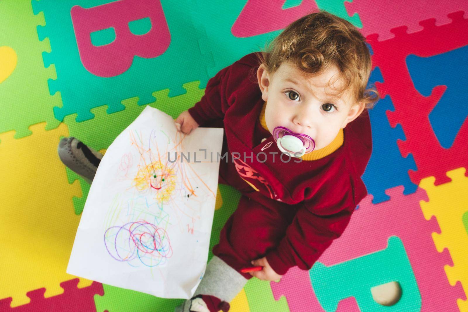 Cute baby girl with dummy on alphabet play mat drawing on paper with a colored crayon