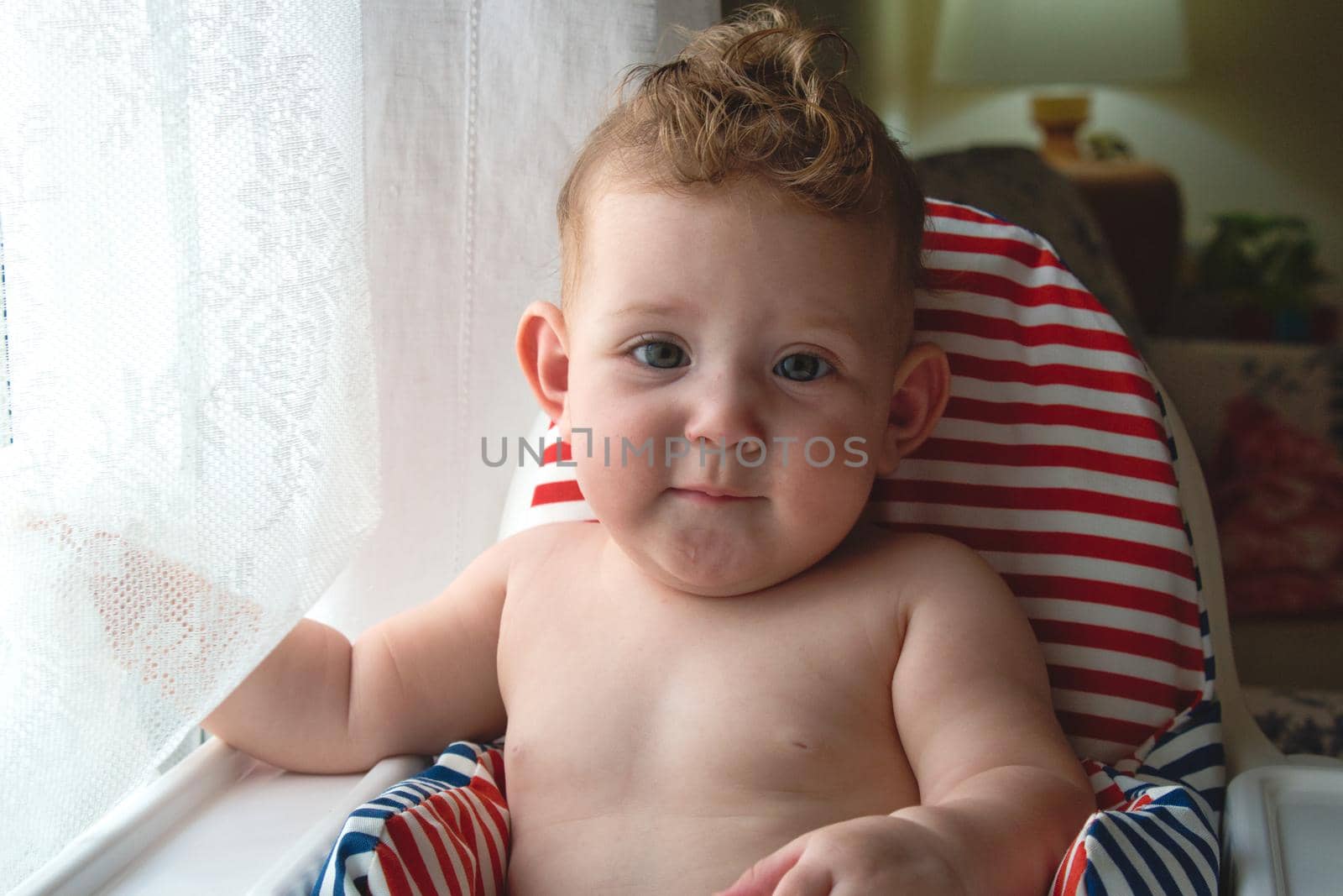 Beautiful caucasian baby girl sitting in a high-chair by a window looking at the camera by tennesseewitney