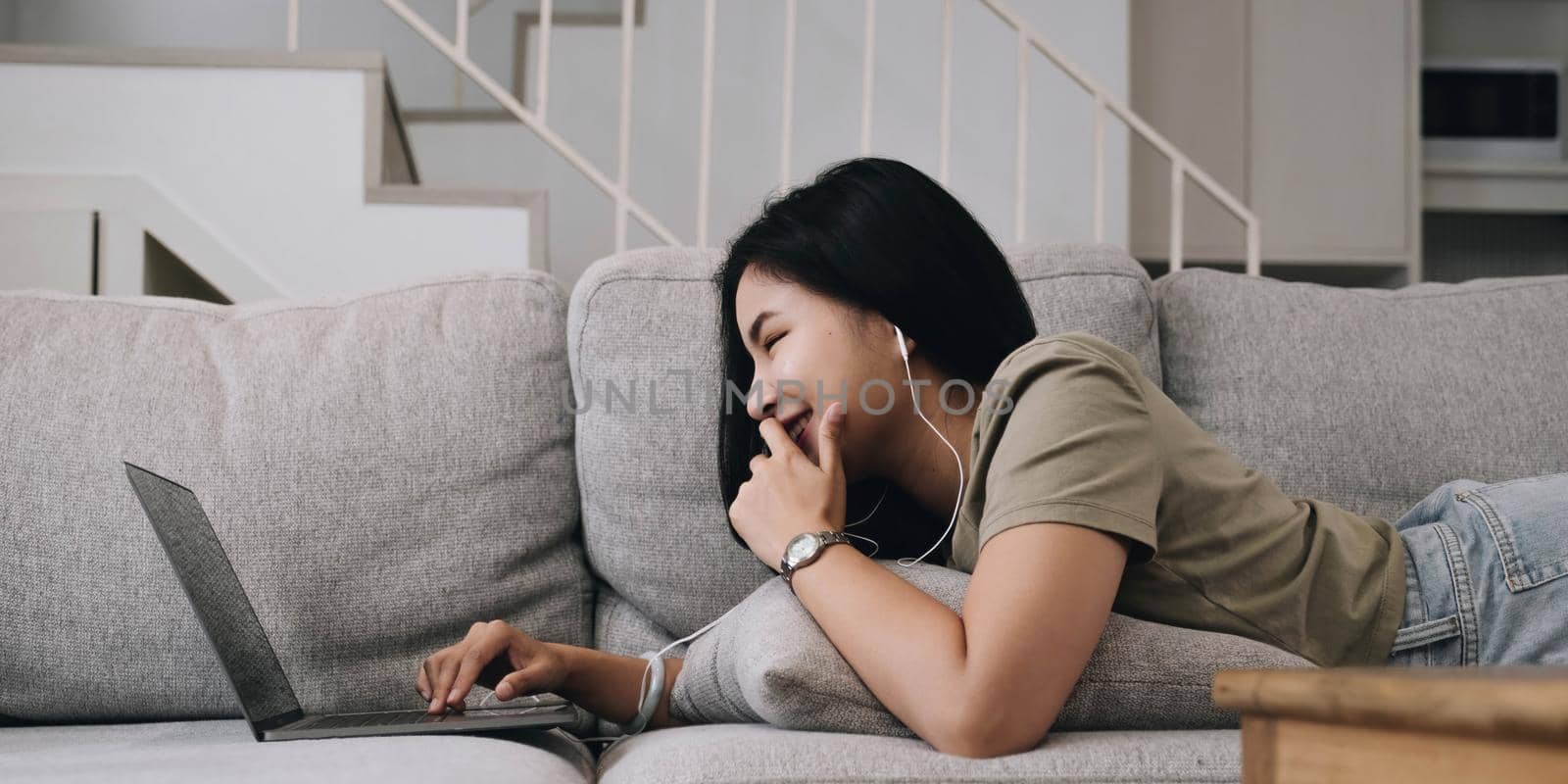 Asian woman lying on the sofa in the living room listening to music on a laptop device woman relaxing on the sofa at home View messages on the computer screen on the device. communication concept.