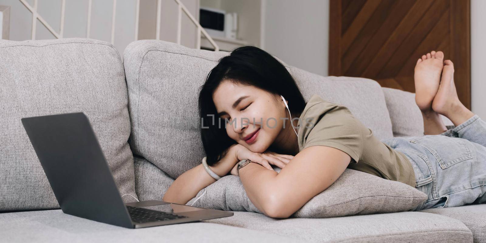 Asian woman lying on the sofa in the living room with her eyes closed, listening to music on a laptop device. woman relaxing on the sofa at home View messages on the computer screen on the device. communication concept by wichayada