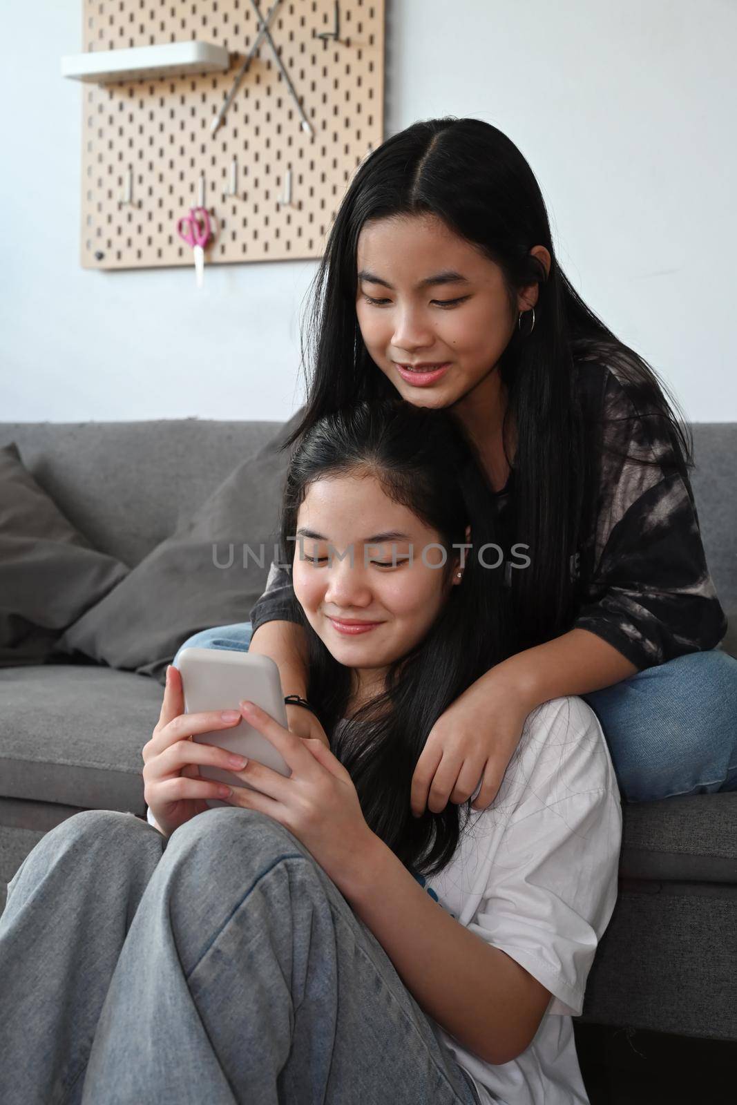 Two cheerful teenage girls sitting in living room and using smart phone.
