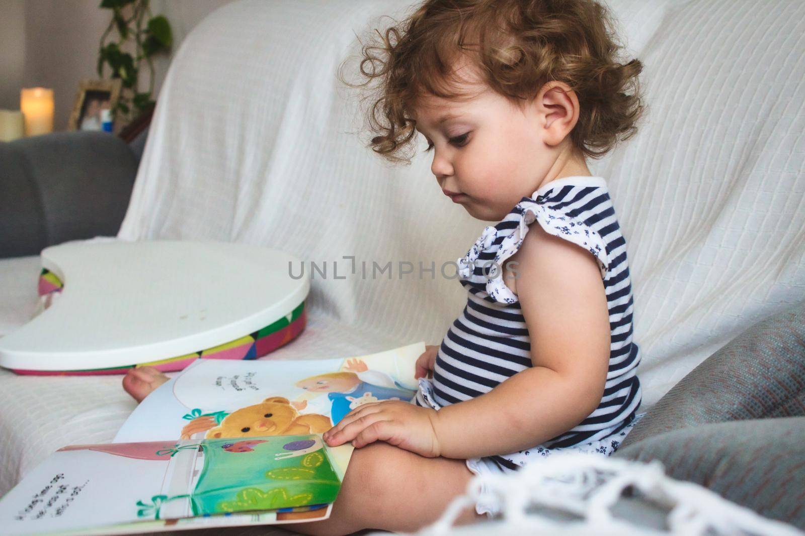 Little baby girl reading a picture book on a white sofa by tennesseewitney