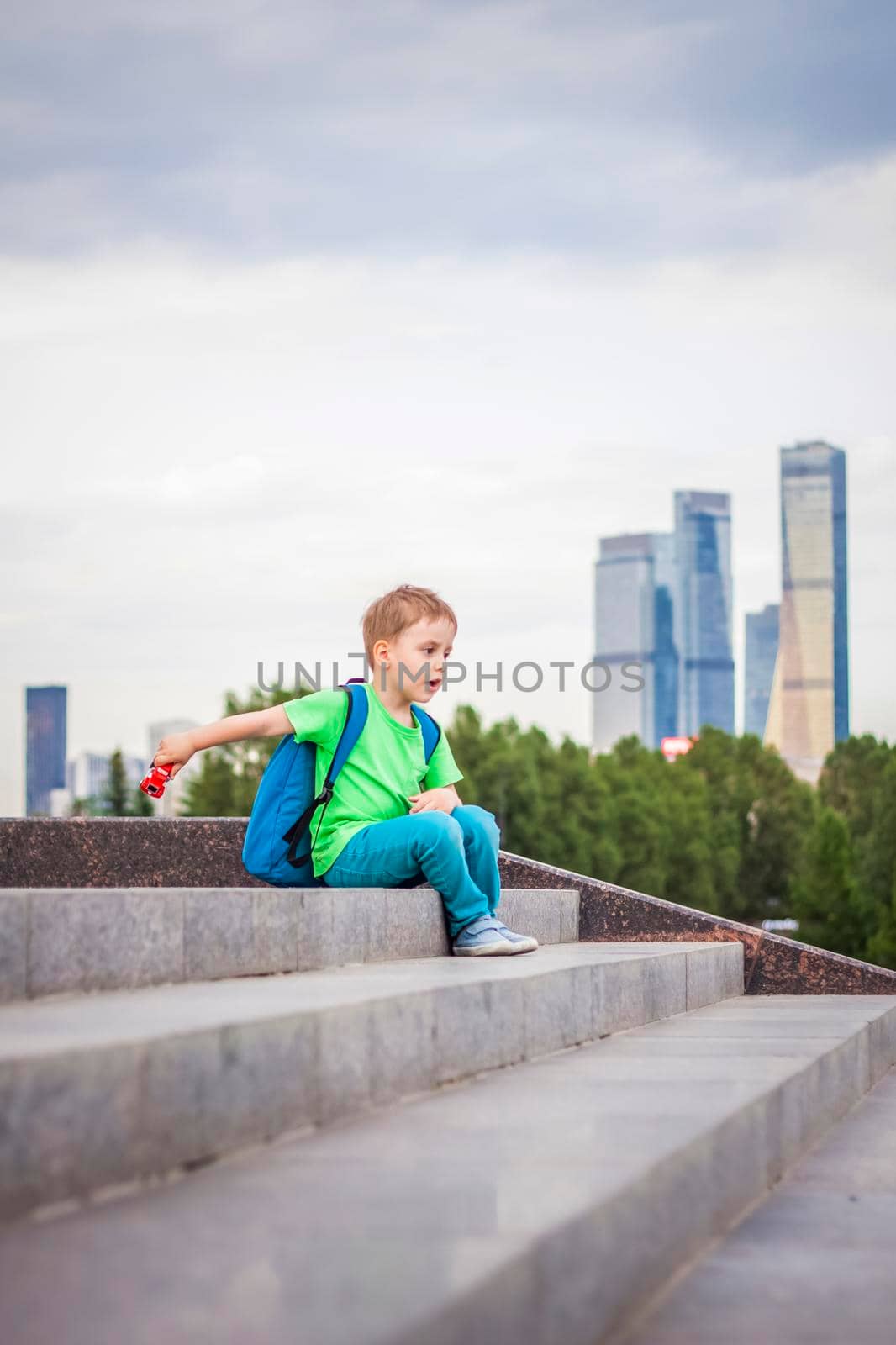 A boy is playing with a toy, sitting on the steps in the open air against the backdrop of skyscrapers and high-rise buildings. Journey. Lifestyle in the city. Center, streets.