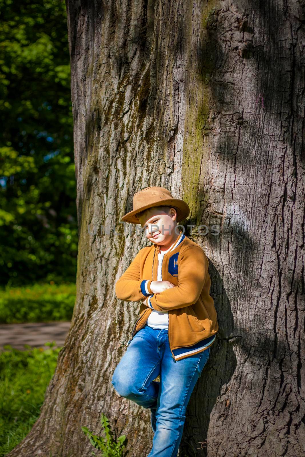 Cute boy posing in a cowboy hat in the woods by a tree. The sun's rays envelop the space. Interaction history for the book. Space for copying. Selective focus.