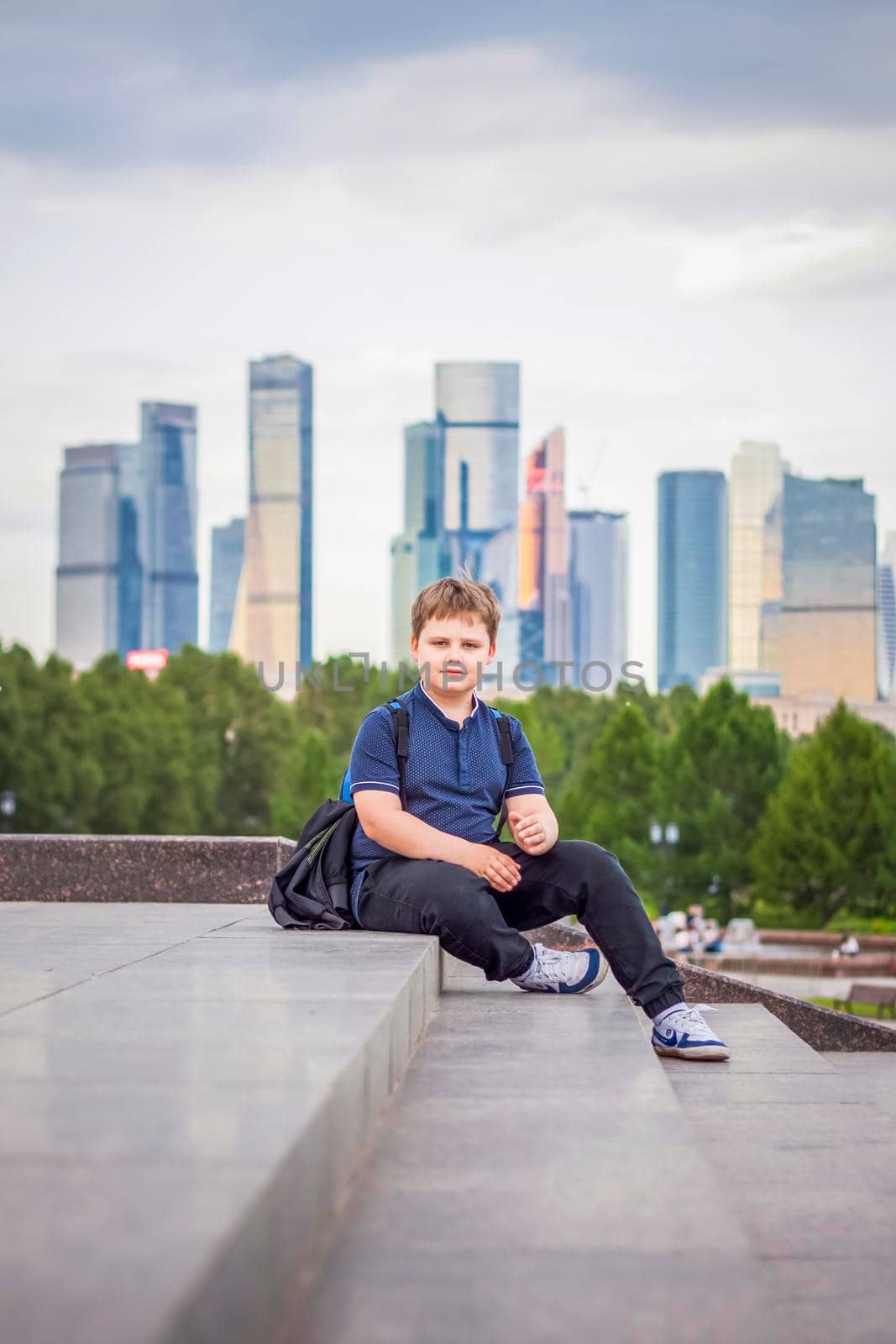 The boy is sitting on the steps in the open air against the background of skyscrapers and high-rise buildings. Journey. Lifestyle in the city. Center, streets.