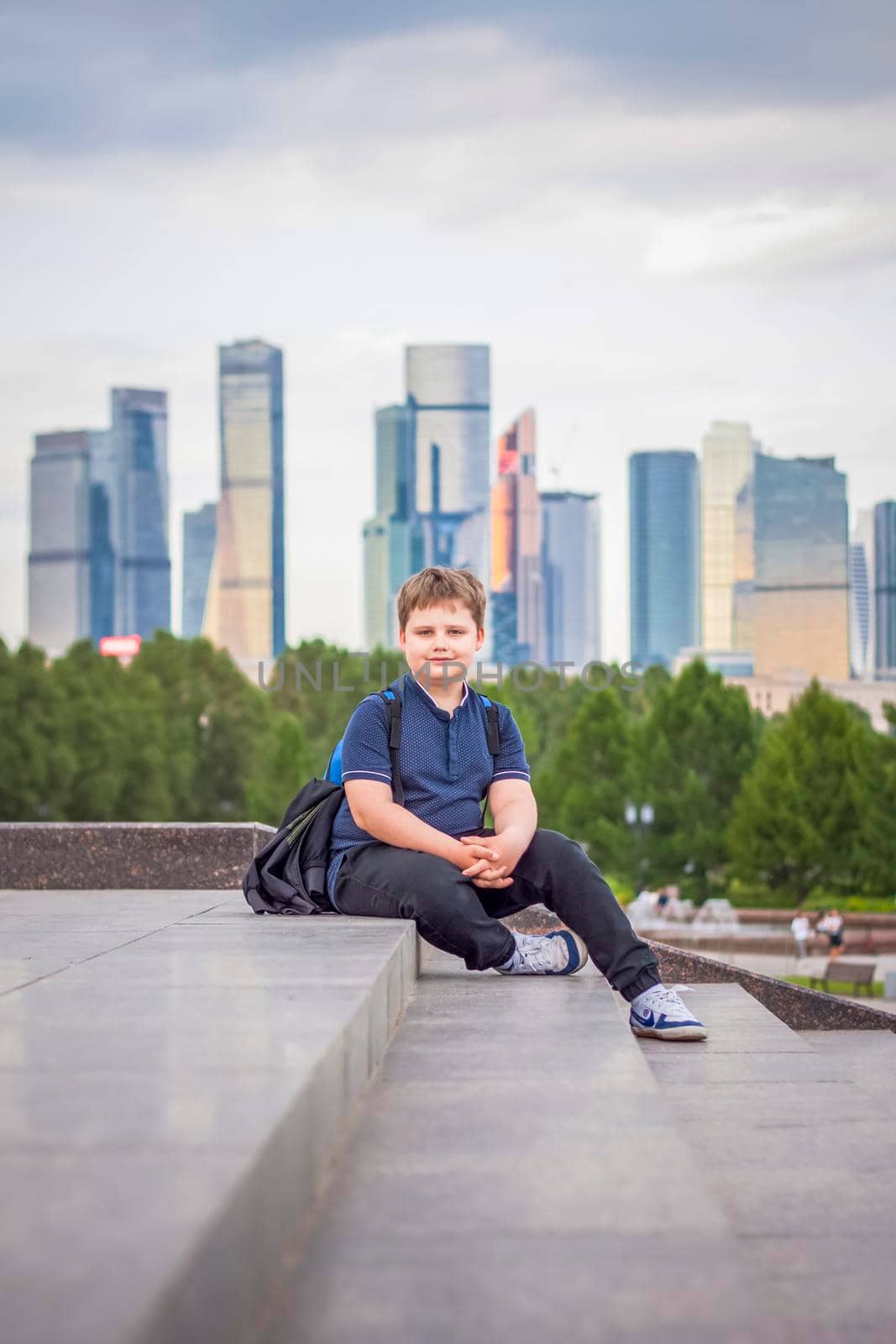 The boy is sitting on the steps in the open air against the background of skyscrapers and high-rise buildings. Journey. Lifestyle in the city. Center, streets.