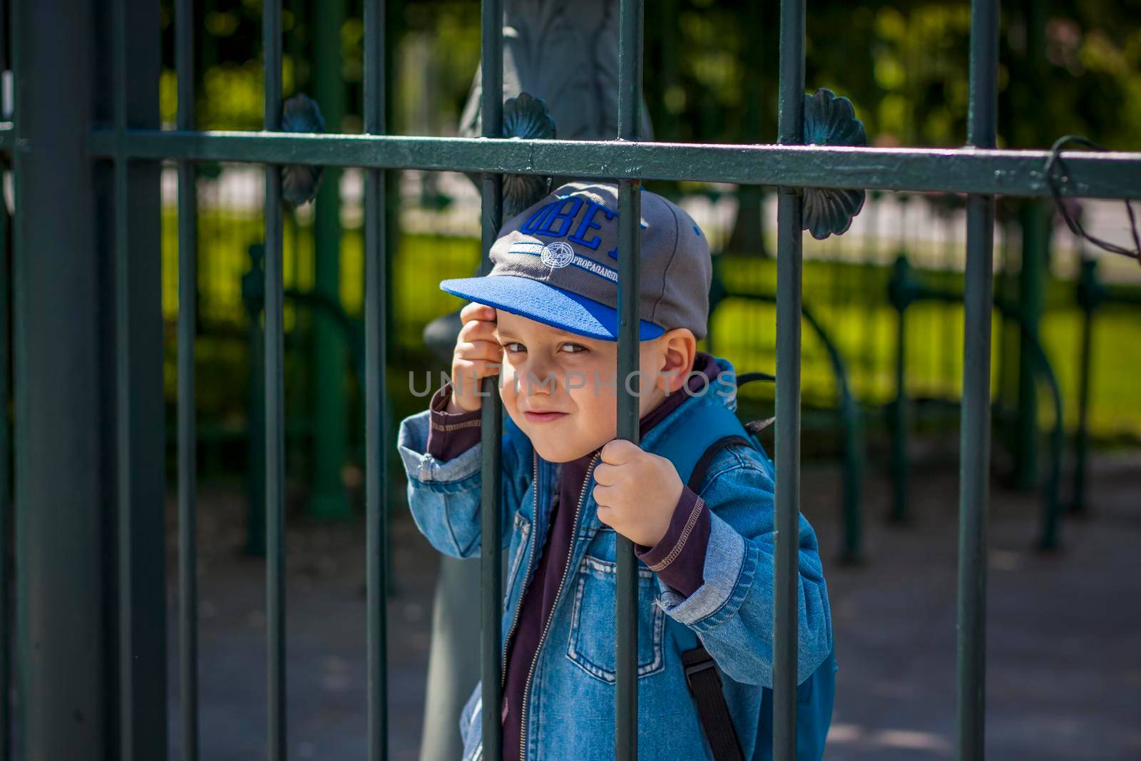 A boy in a baseball cap looks through the iron bars of the fence.  Interactions. Selective focus. by Alina_Lebed