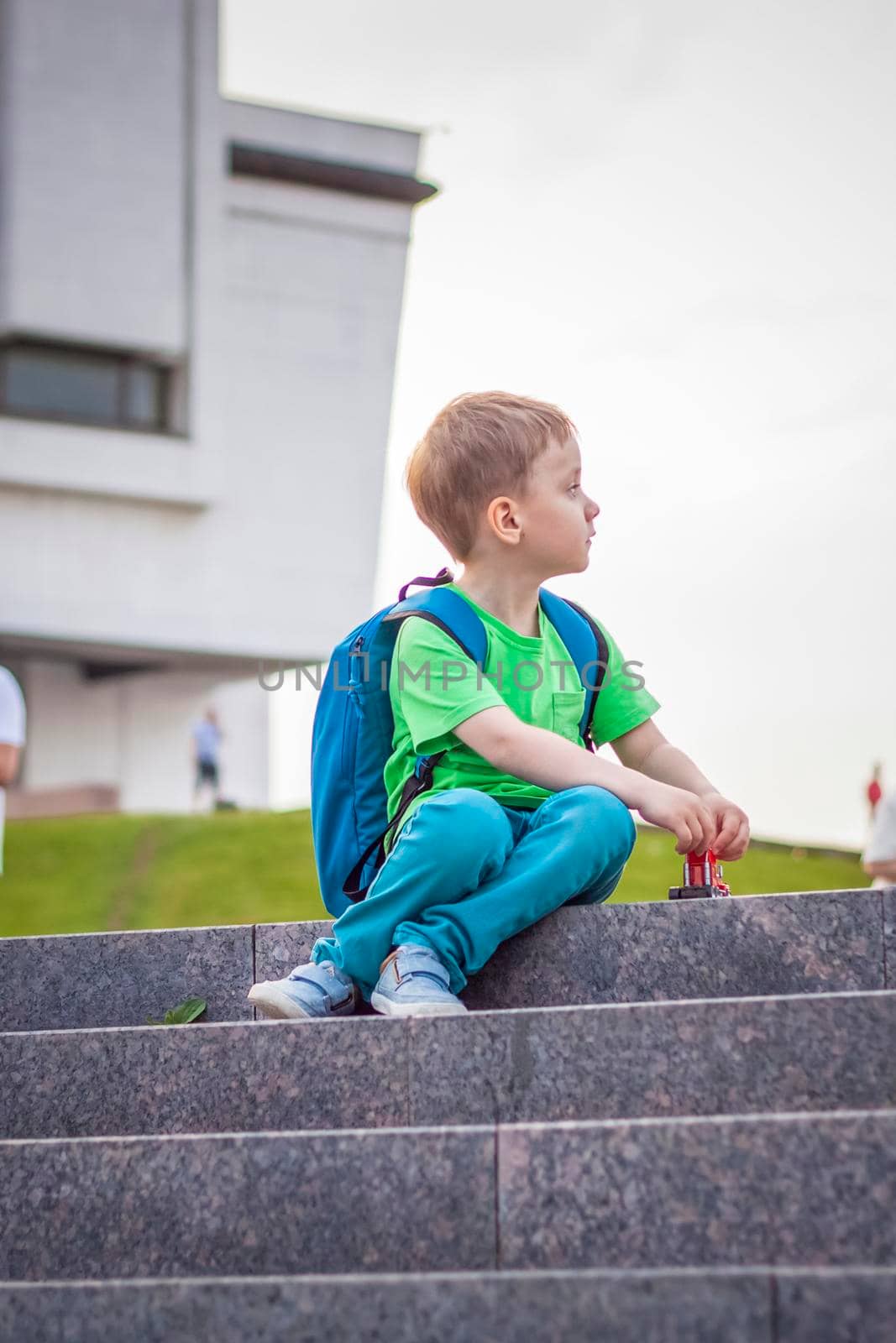 A boy is playing with a toy, sitting on the steps in the open air against the backdrop of skyscrapers and high-rise buildings. Journey. Lifestyle in the city. Center, streets.