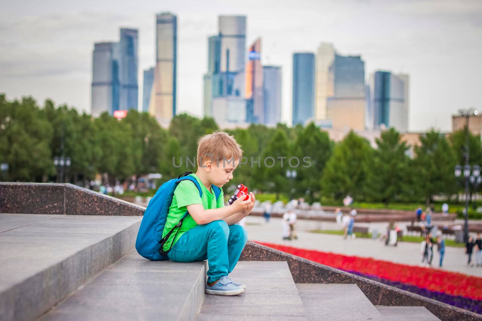 A boy is playing with a toy, sitting on the steps in the open air against the backdrop of skyscrapers and high-rise buildings. Journey. Lifestyle in the city. Center, streets.