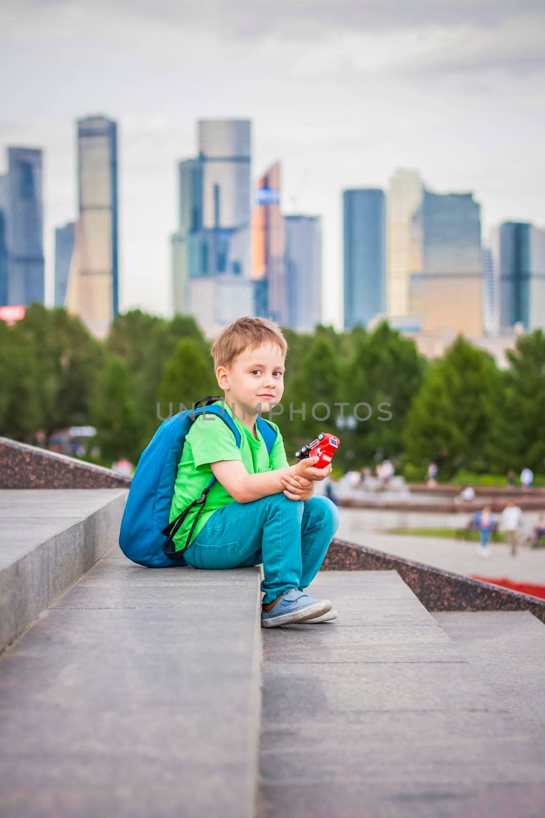 A boy is playing with a toy, sitting on the steps in the open air against the backdrop of skyscrapers and high-rise buildings. Journey. Lifestyle in the city. Center, streets.