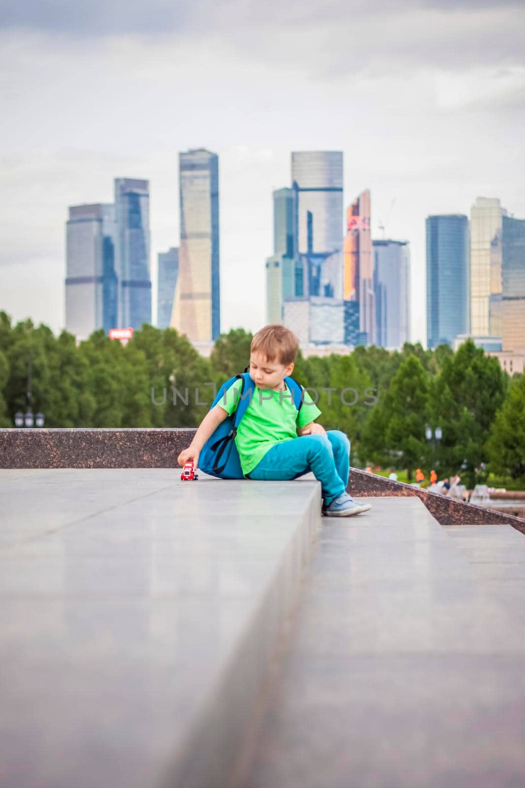 A boy is playing with a toy, sitting on the steps in the open air against the backdrop of skyscrapers and high-rise buildings. Journey. Lifestyle in the city. Center, streets.