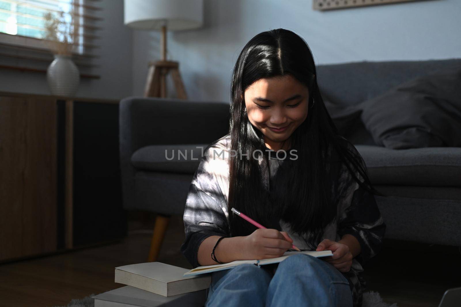 Smiling young asian girl sitting on floor in living room and doing homework.