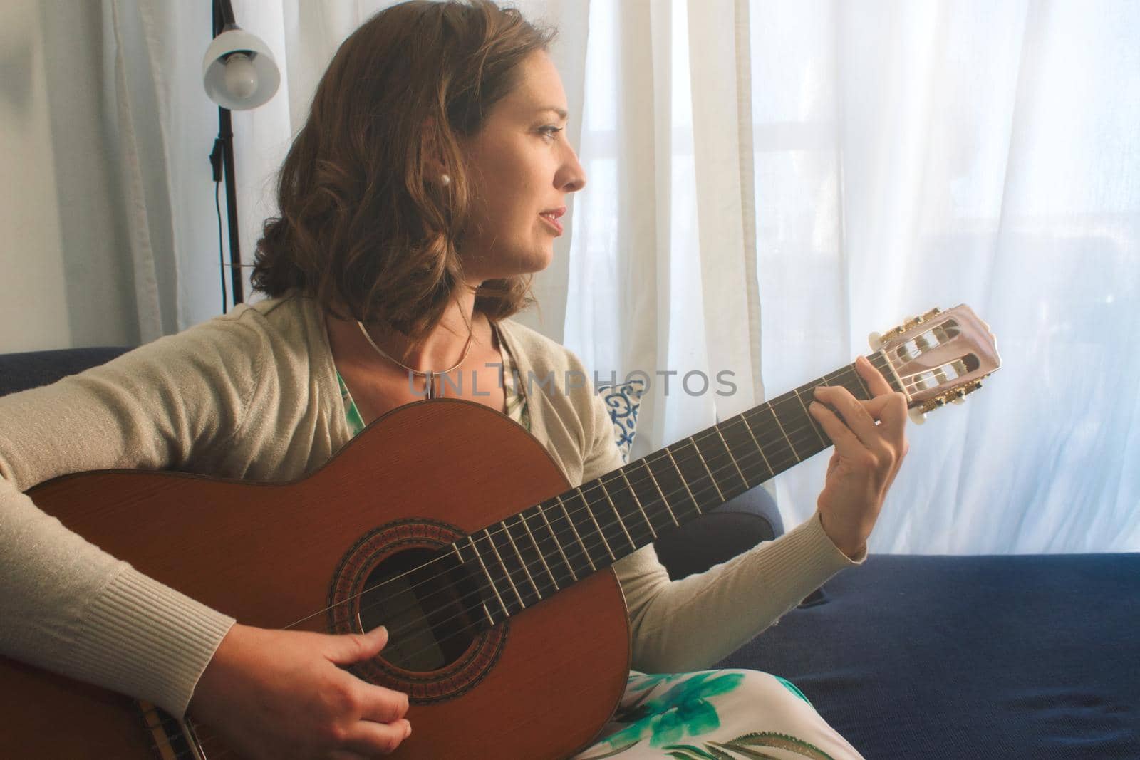 Attractive young woman sitting on a sofa at home playing the Spanish guitar by tennesseewitney